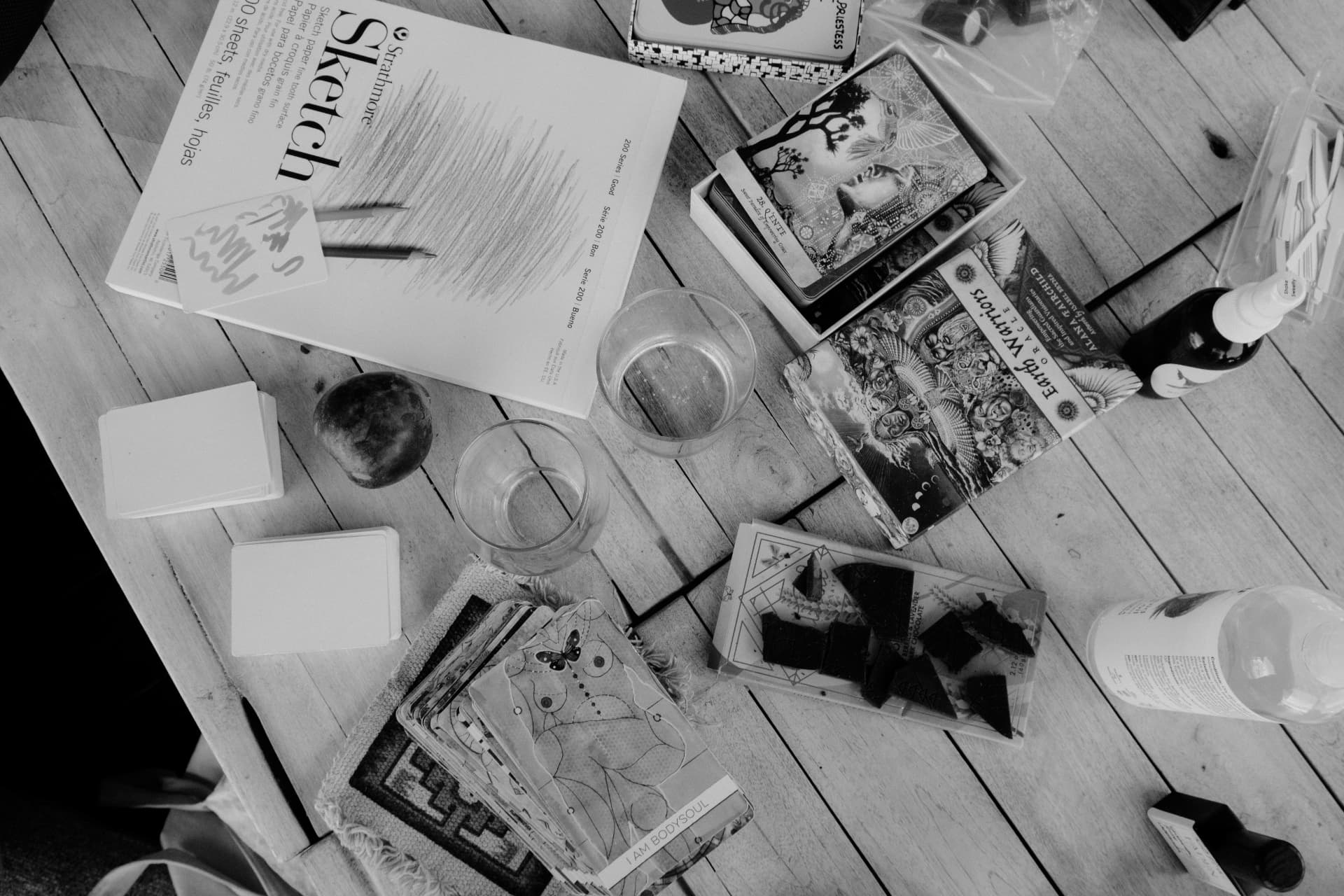An overhead view of sketchbooks, tarot cards and glasses on a wooden floor, in black and white