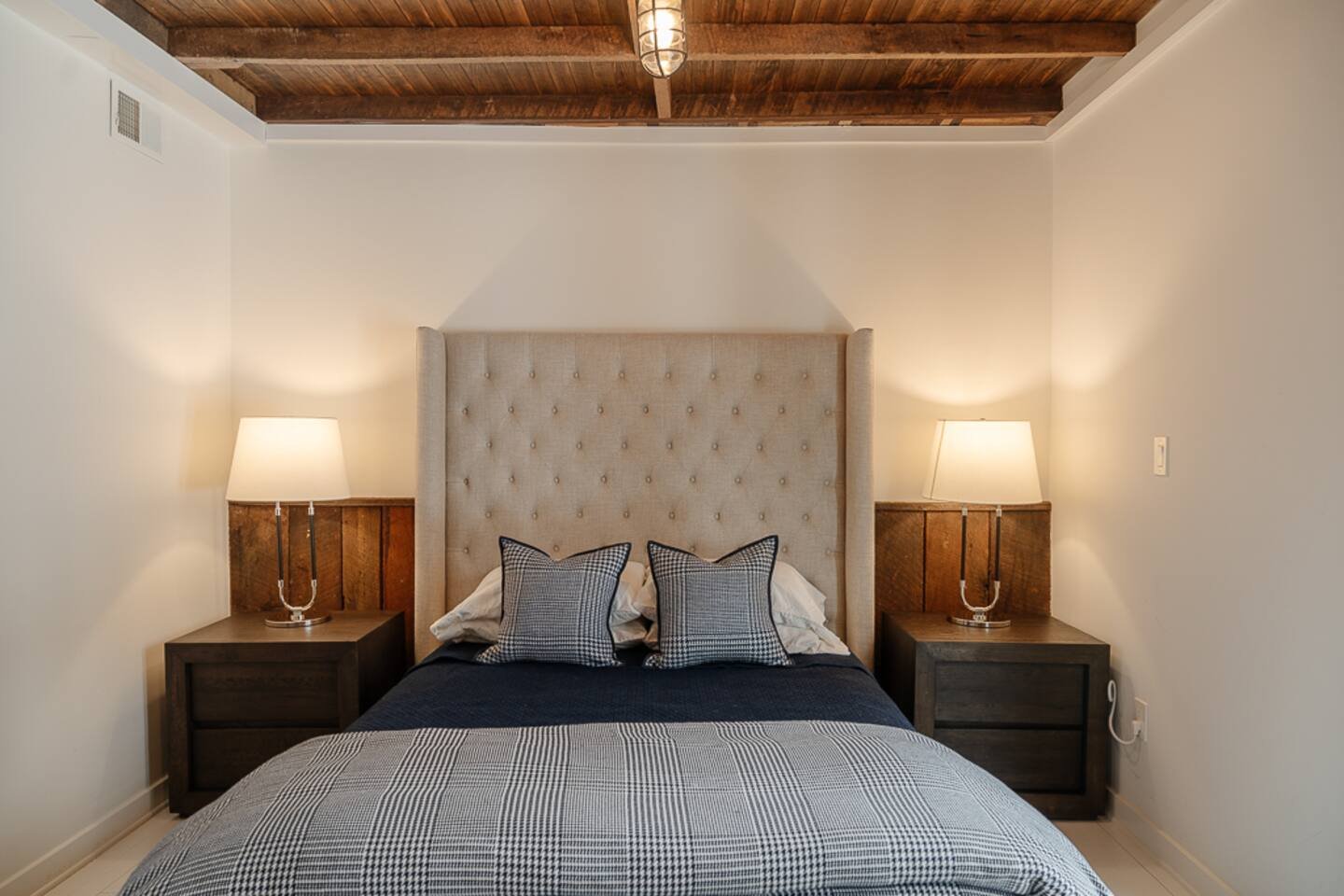 A bedroom with a tall upholstered headboard and exposed wooden ceiling beams