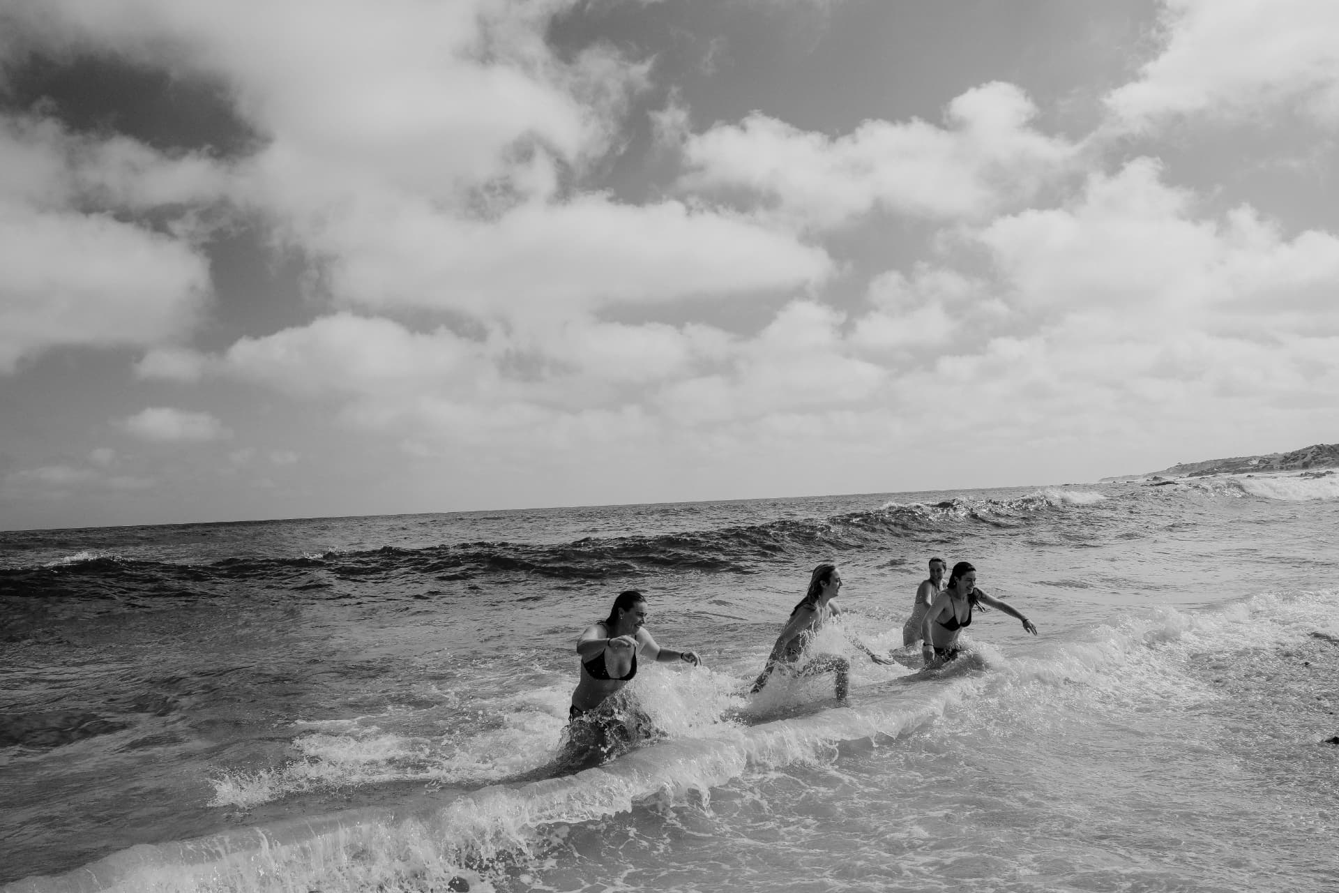 Four women playing together in breaking ocean waves beneath a cloud-filled sky, in black and white