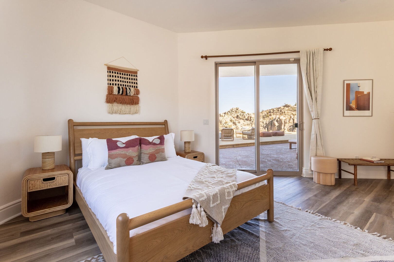 A bedroom with a wooden queen bed and a glass door opening to desert boulders