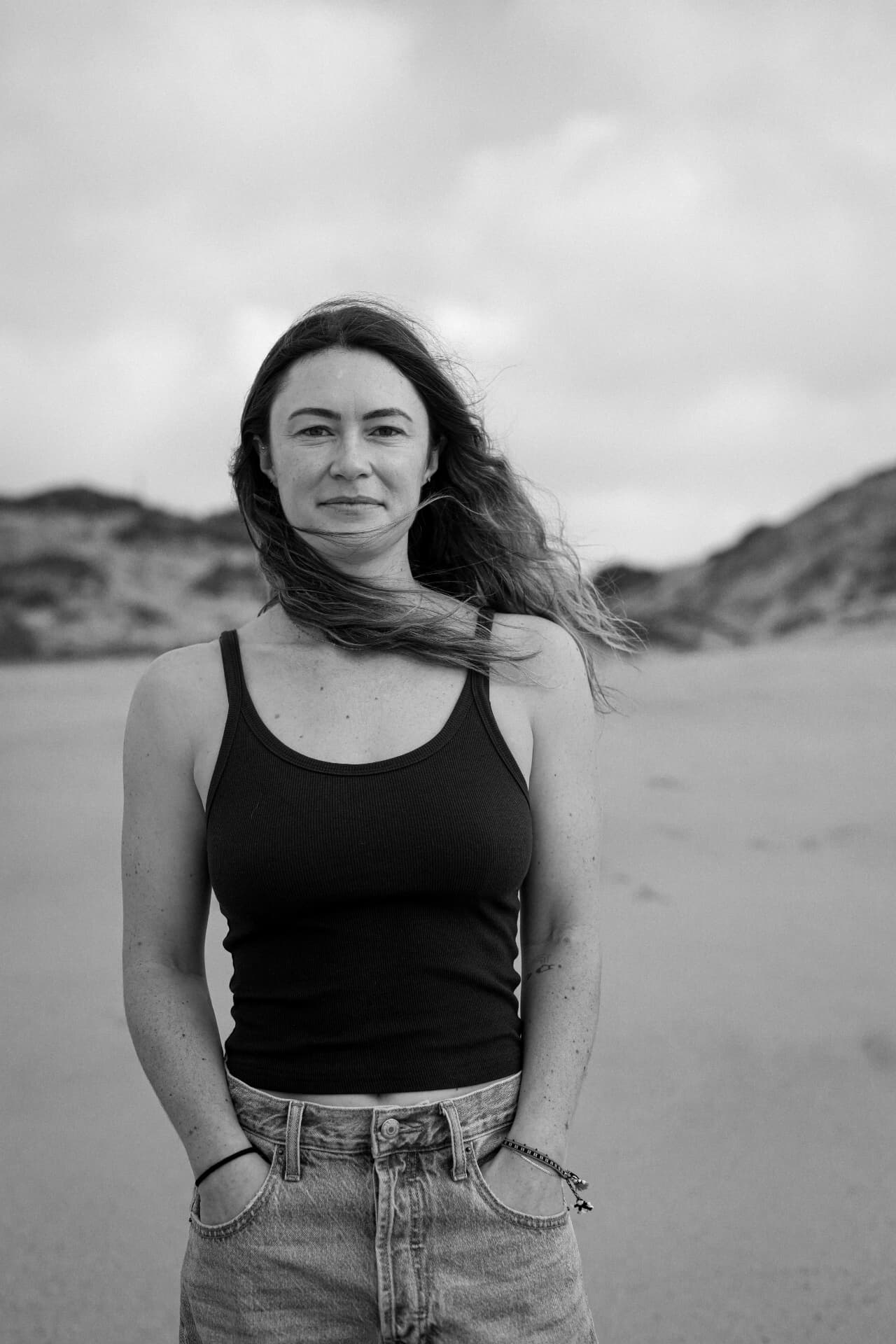 A woman standing on the beach with her hands in her pockets, in black and white