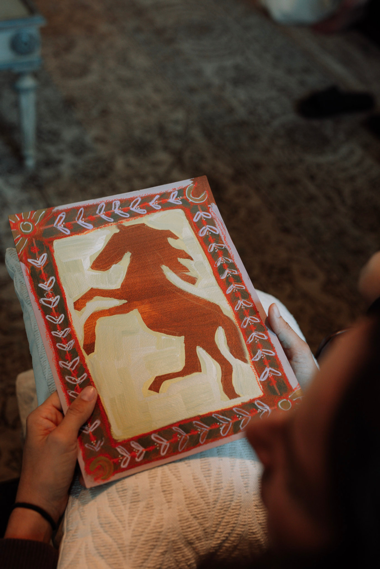 Hands holding a painted horse with a red botanical border