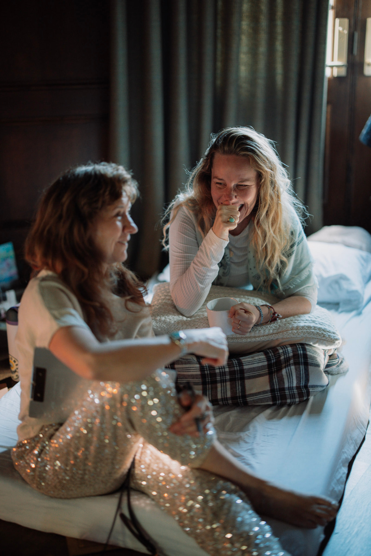 Two women laughing together on a mattress with mugs of tea in morning light