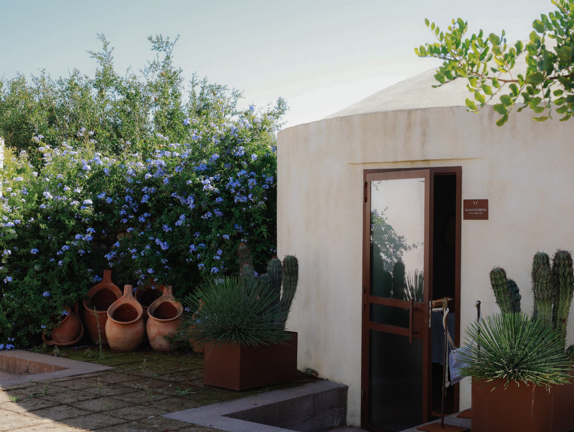 An open doorway in a white rounded building beside flowering plants and terracotta pots