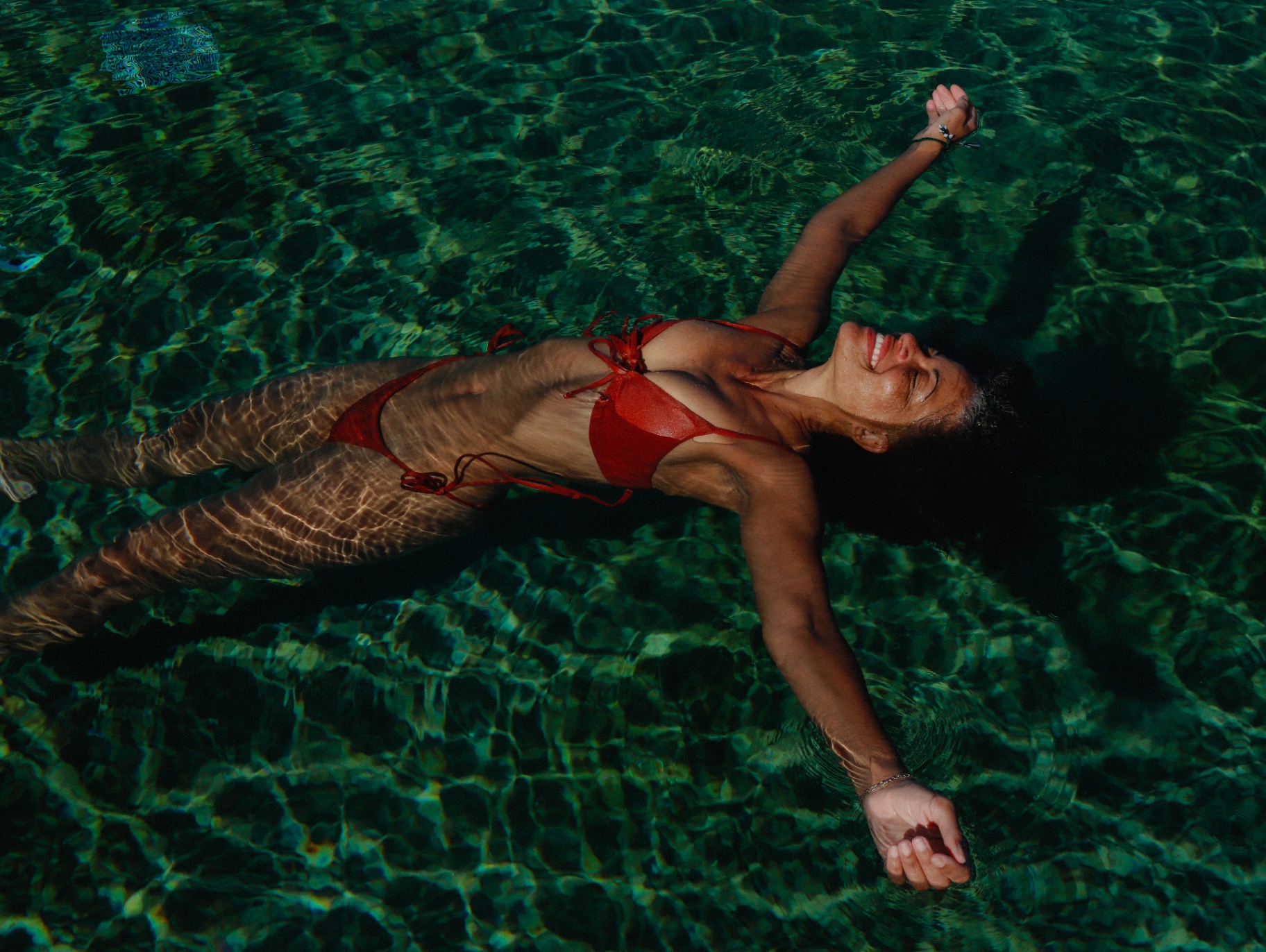 A woman floating with outstretched arms in deep green water, sunlight rippling across her