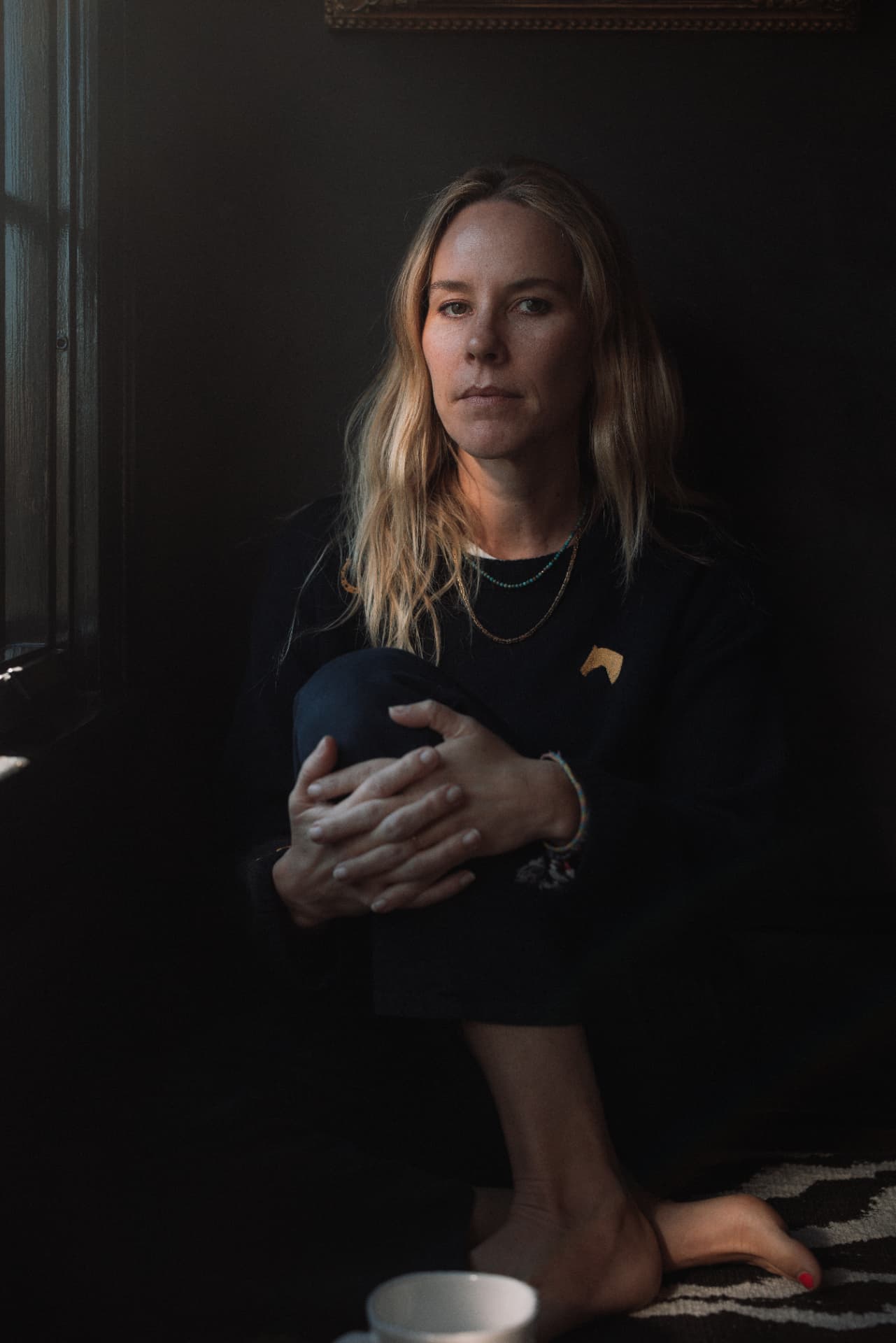 A woman seated beside a window, holding her knee in quiet light