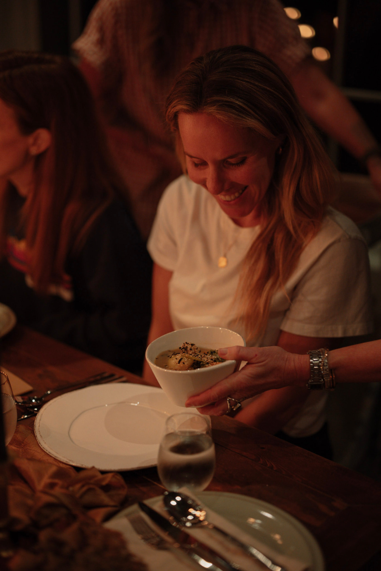 A smiling woman holding a bowl of food at the candlelit dinner table