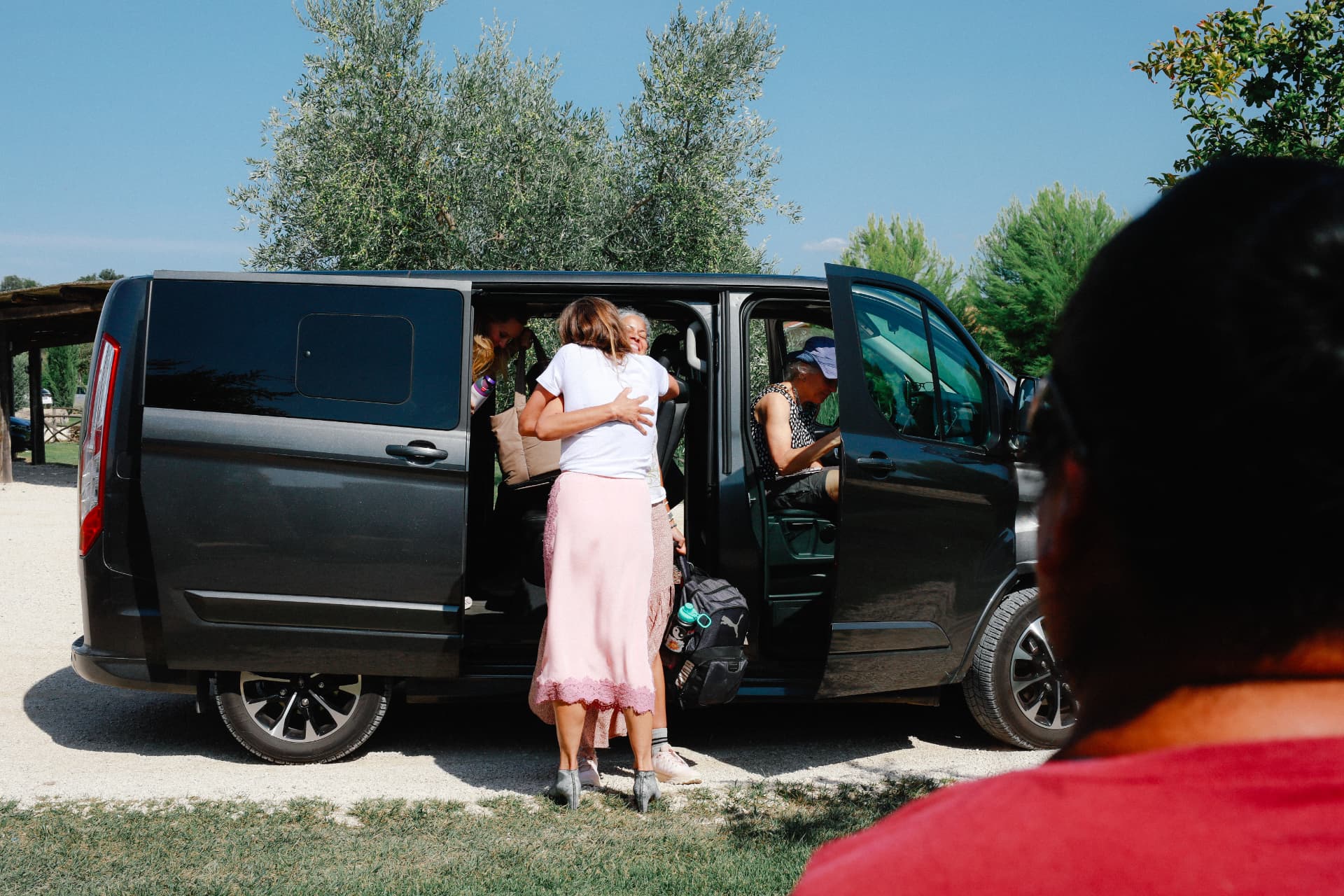 A woman embraced on arrival beside a van beneath olive trees