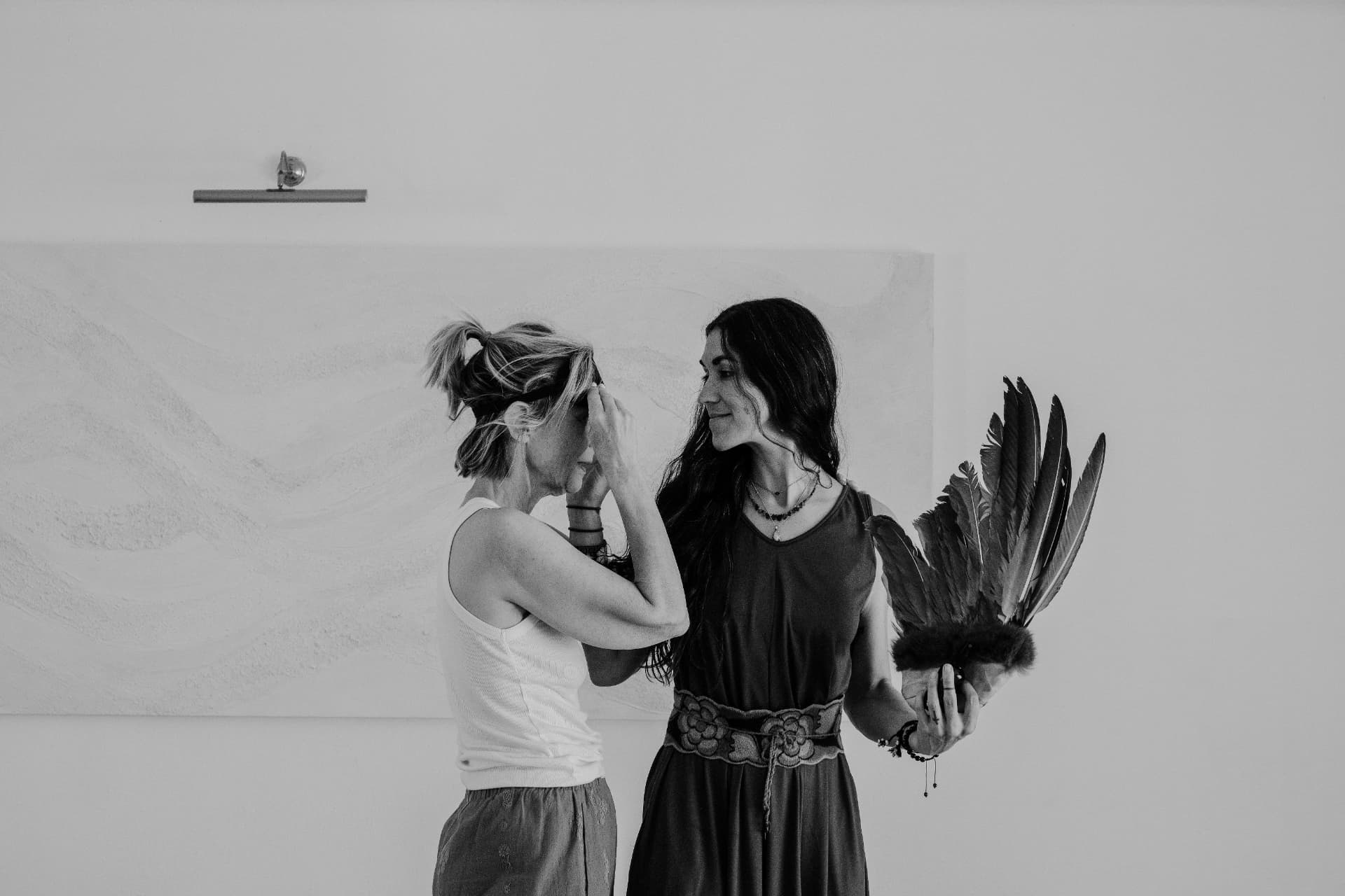 Two women facing one another beside white walls, one holding a feather fan, in black and white