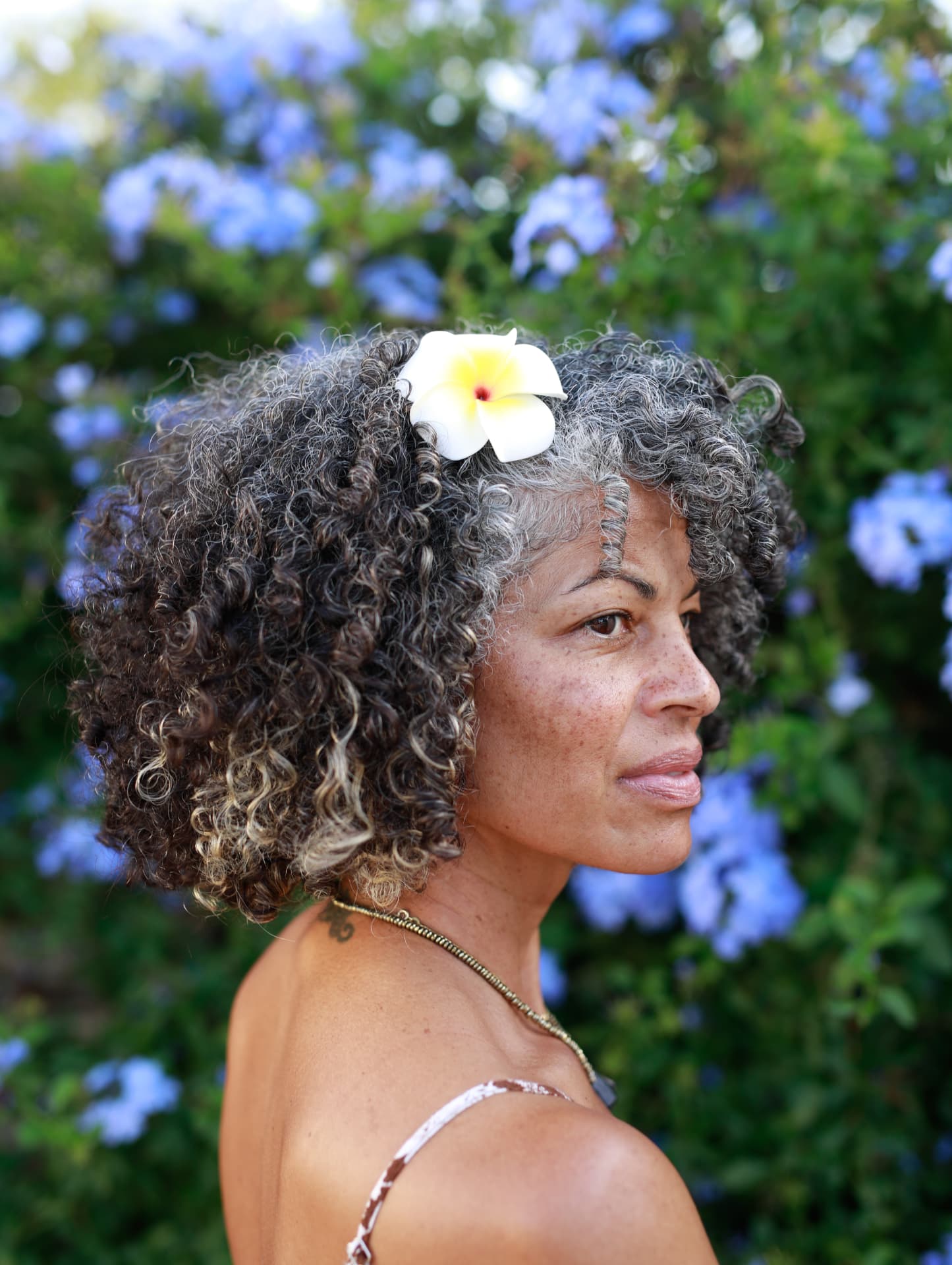 A woman with curly silver hair and a white flower behind her ear before blue plumbago