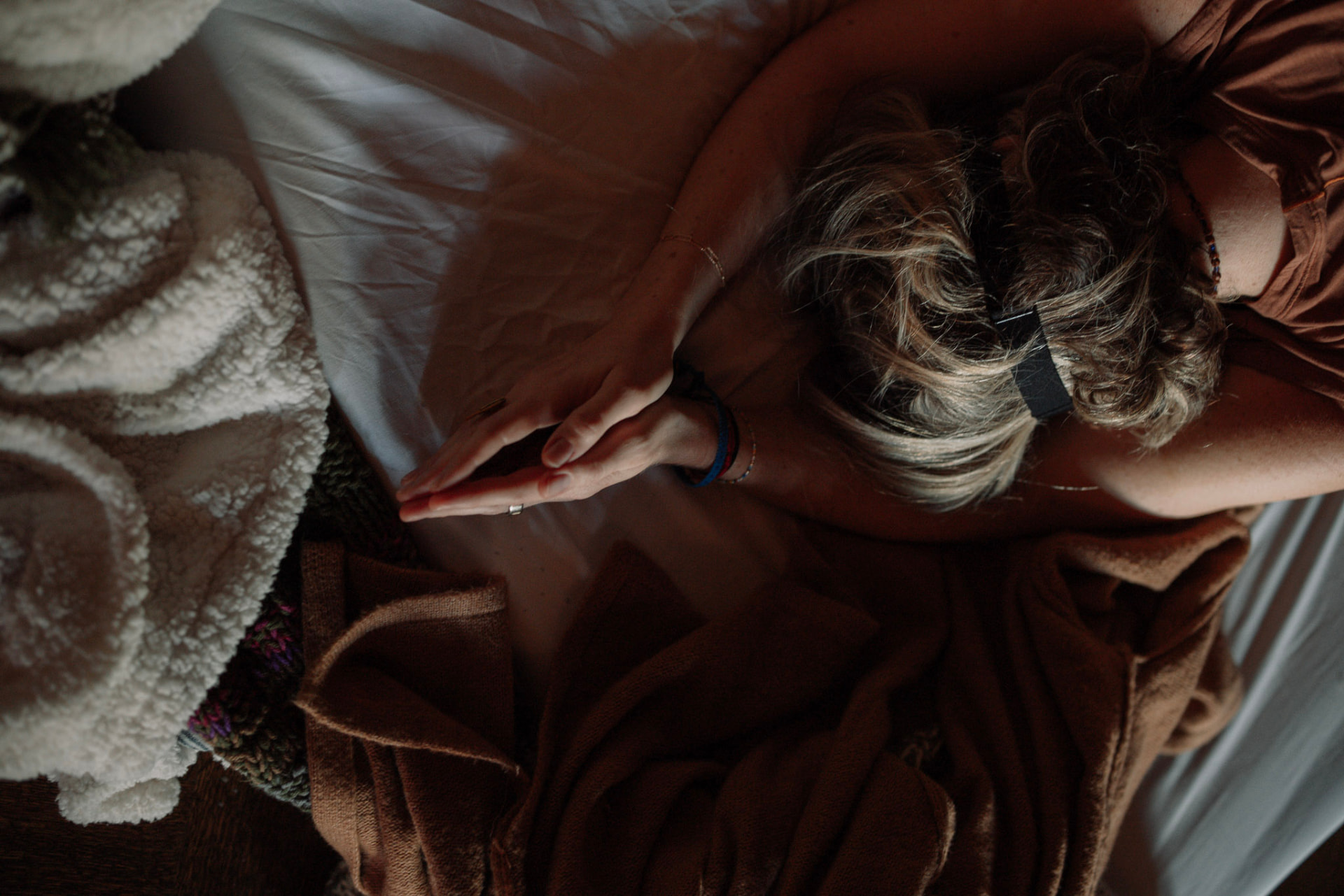 Overhead view of a woman wearing an eye mask, her hands joined above soft blankets