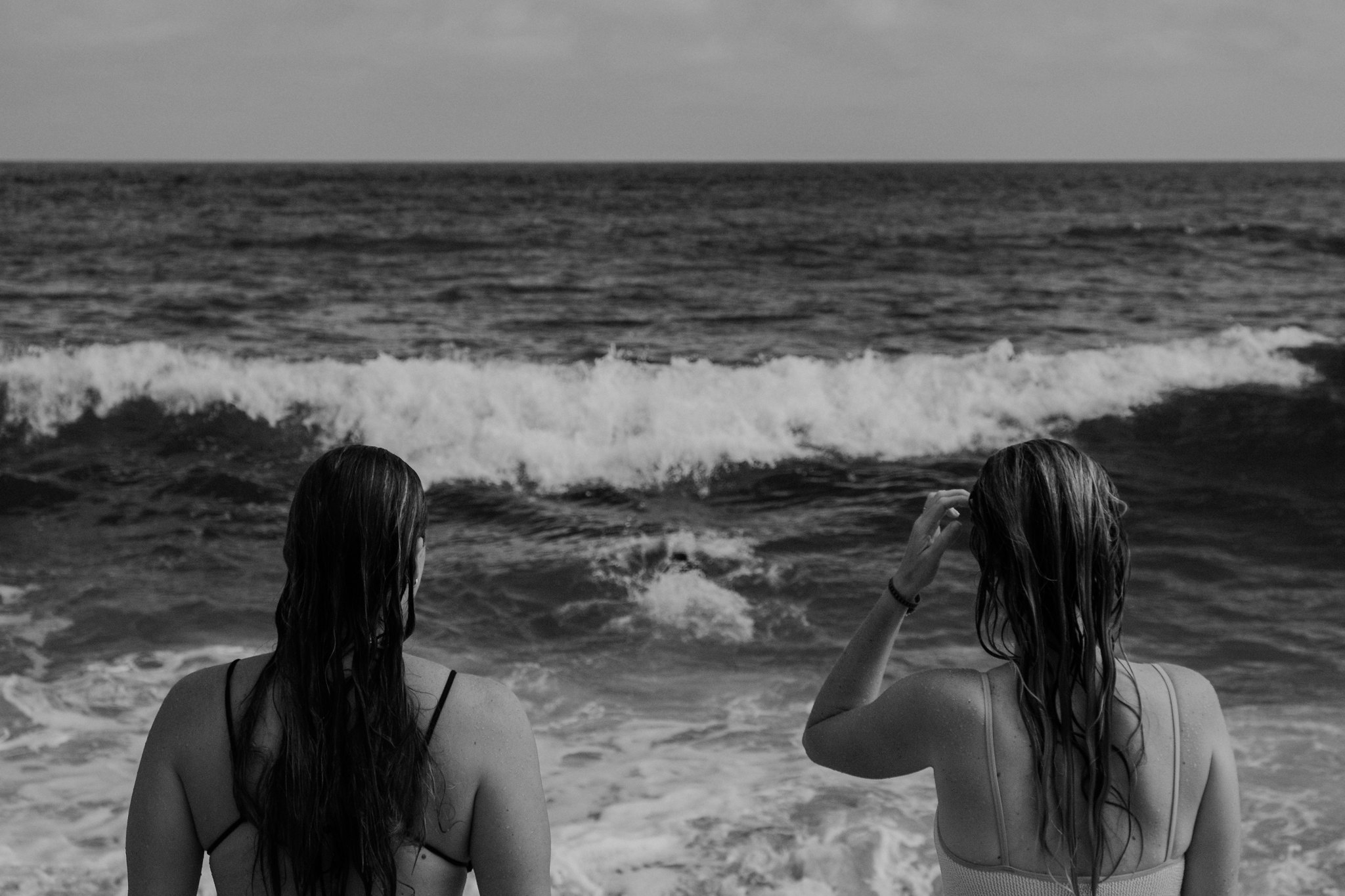 Women standing at the edge of the surf, facing breaking waves, in black and white