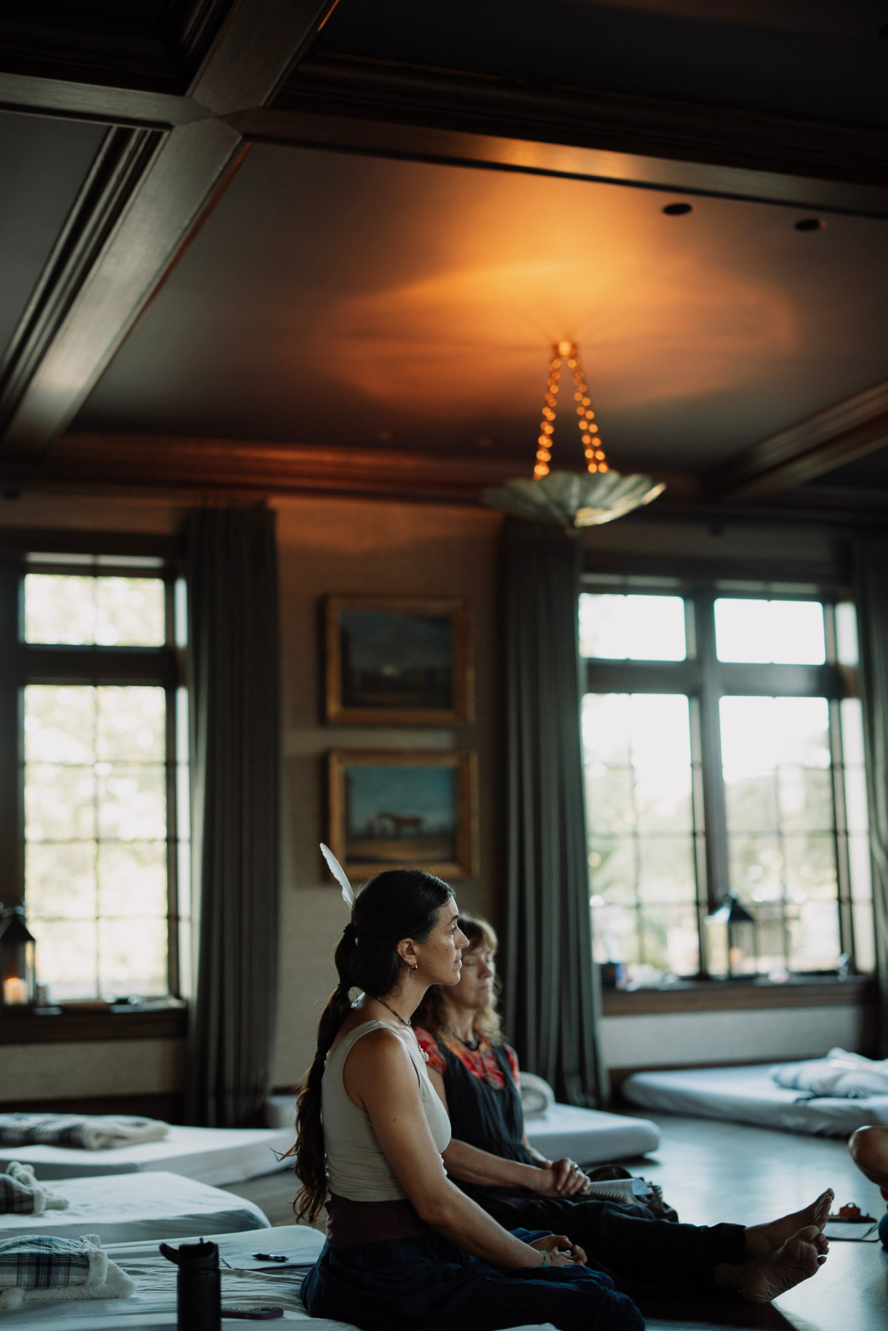 Women seated on mats in a wood panelled room beneath tall windows