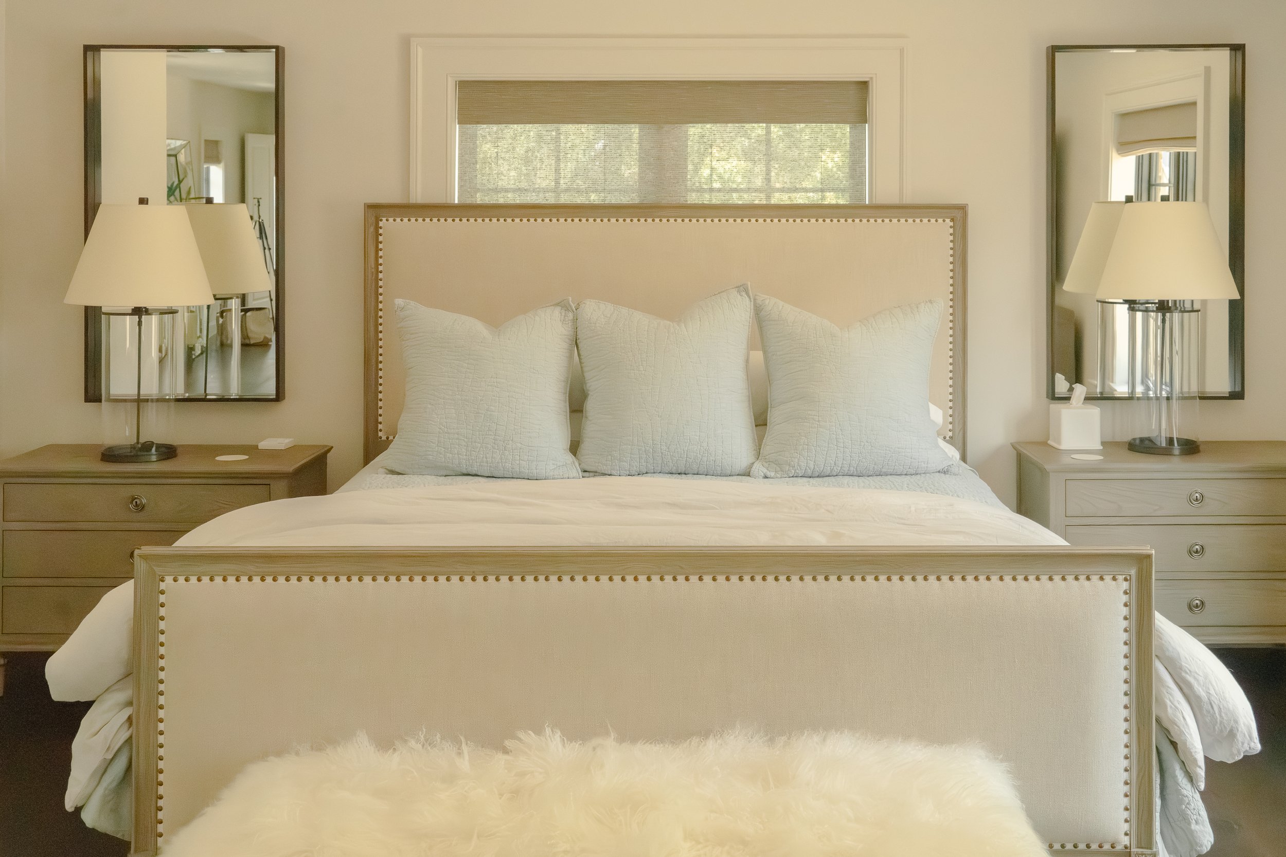 A bedroom with white bedding and a pale upholstered headboard beneath a high window