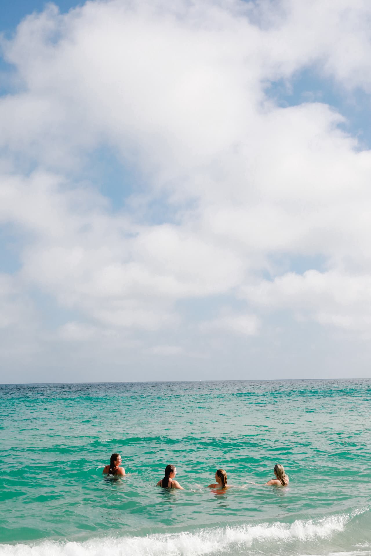 Four women swimming together in the turquoise sea beneath a wide sky