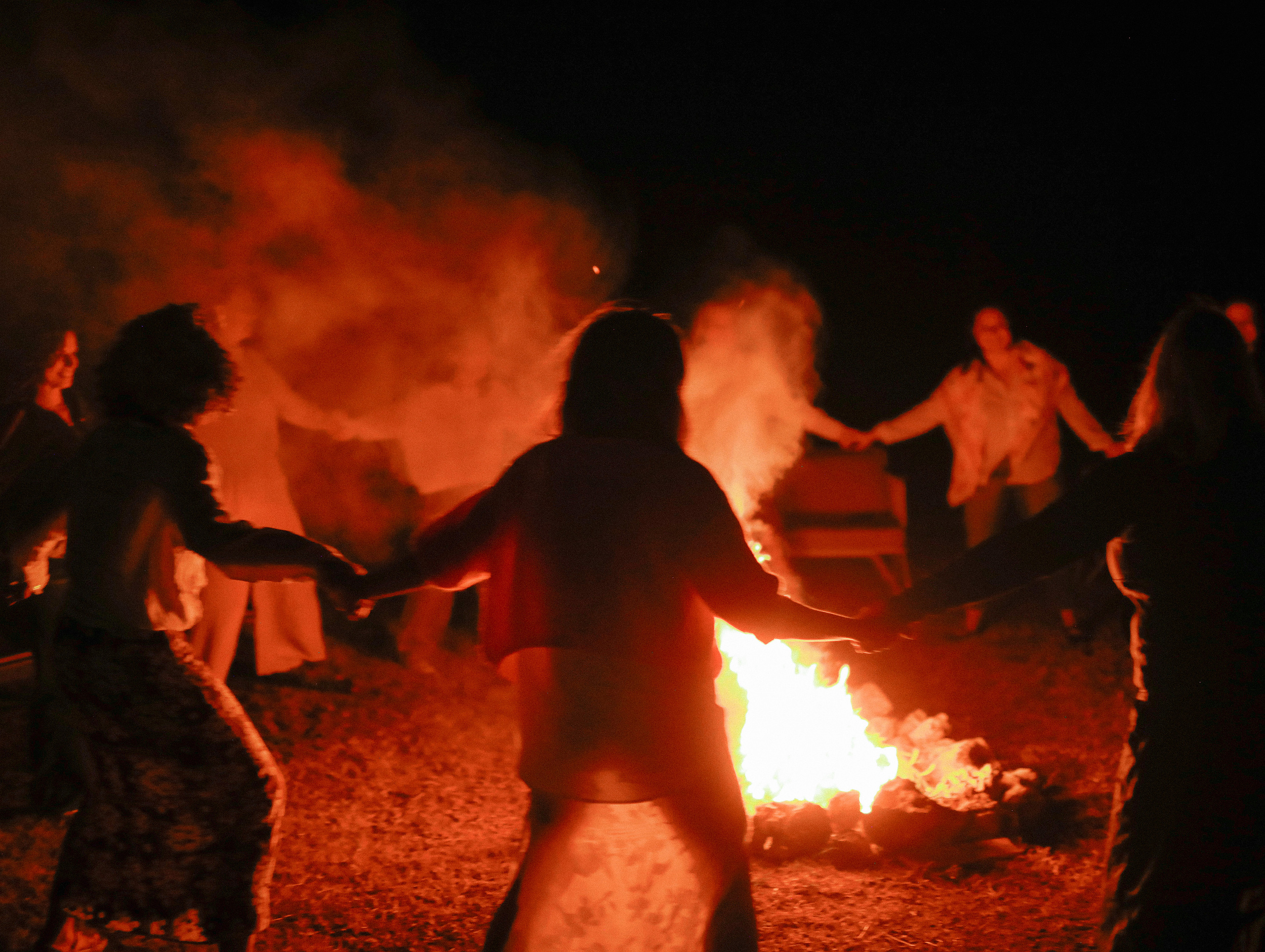 Women holding hands and dancing around a fire at night