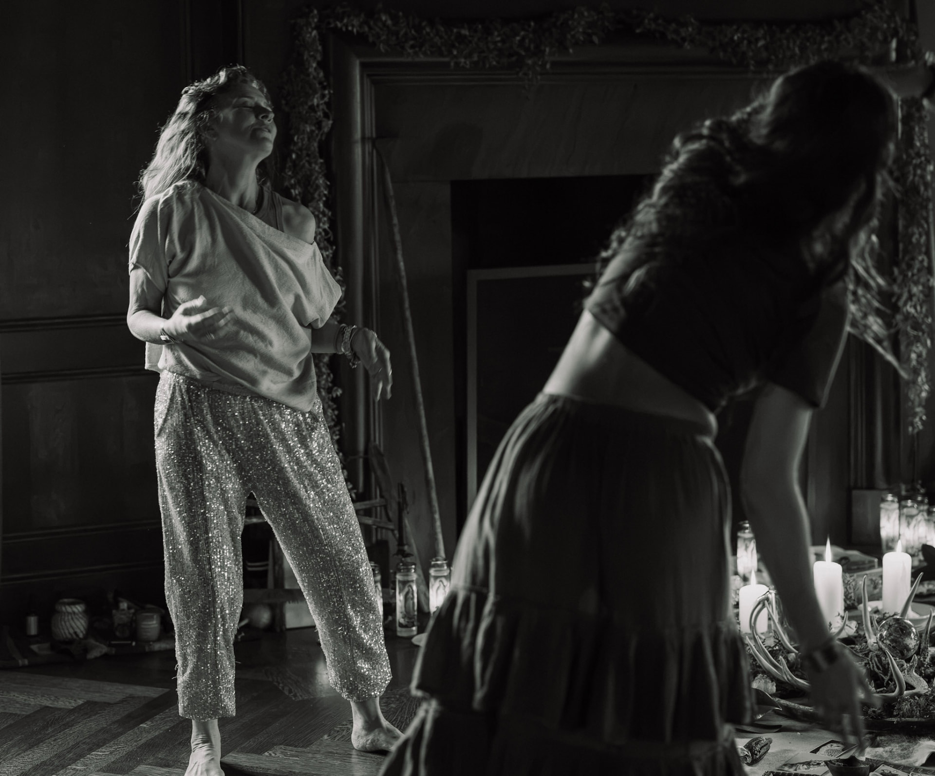 Women moving freely beside a candlelit hearth, in black and white