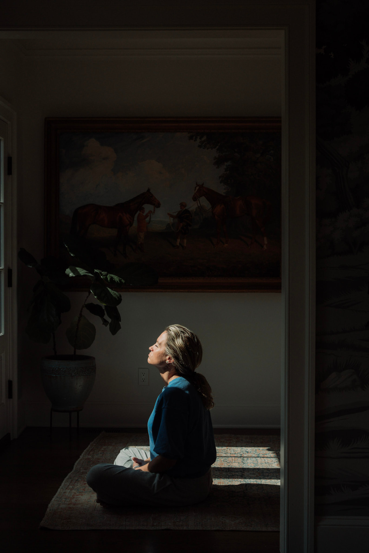 A woman seated cross-legged in a patch of window light beneath a horse painting