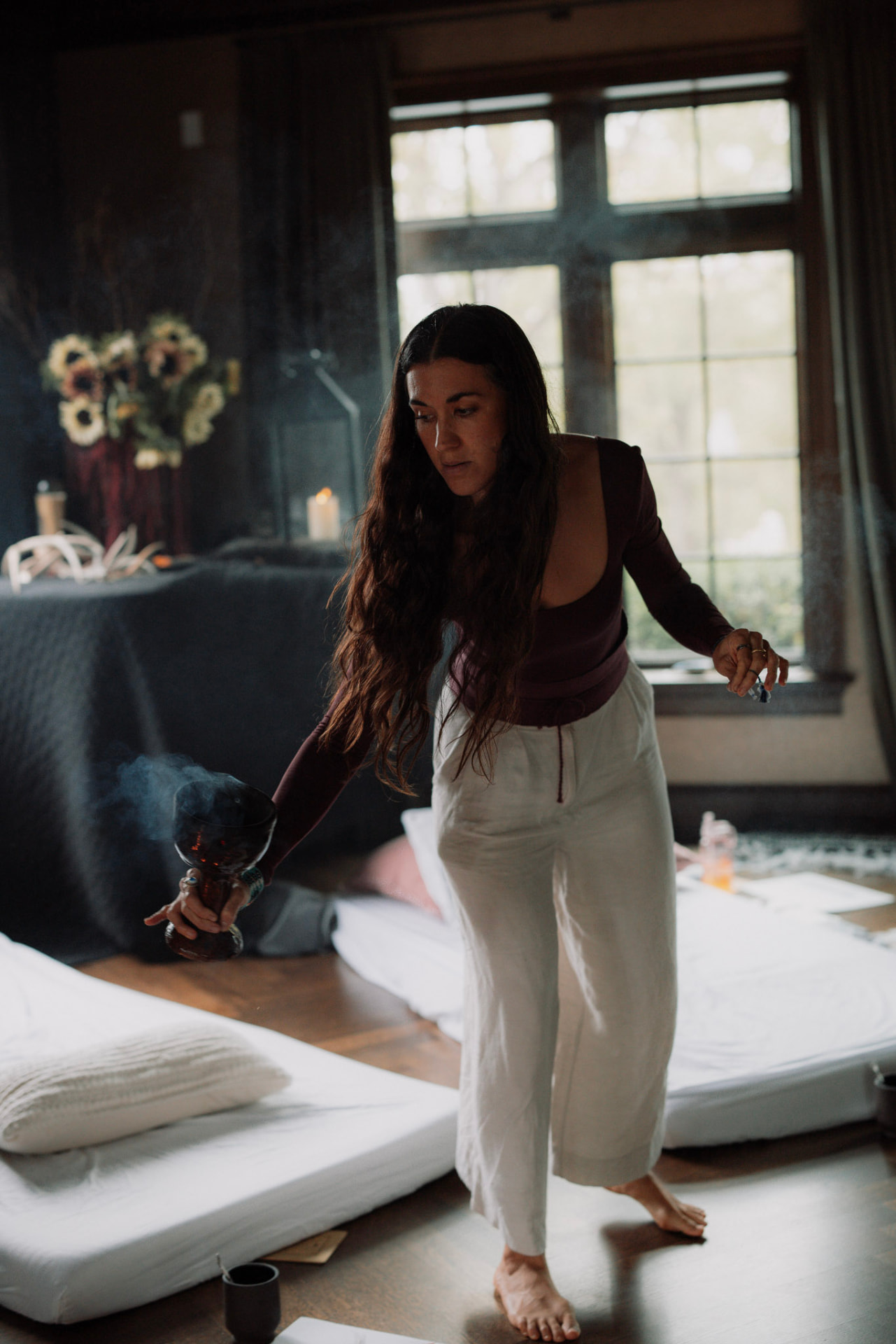A barefoot woman moving slowly between mats beneath tall windows