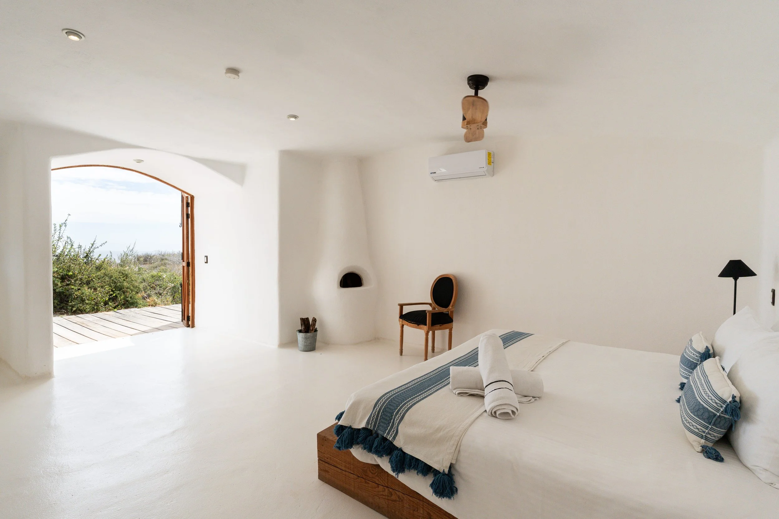 A white bedroom with blue woven bed textiles and an arched doorway opening to a terrace