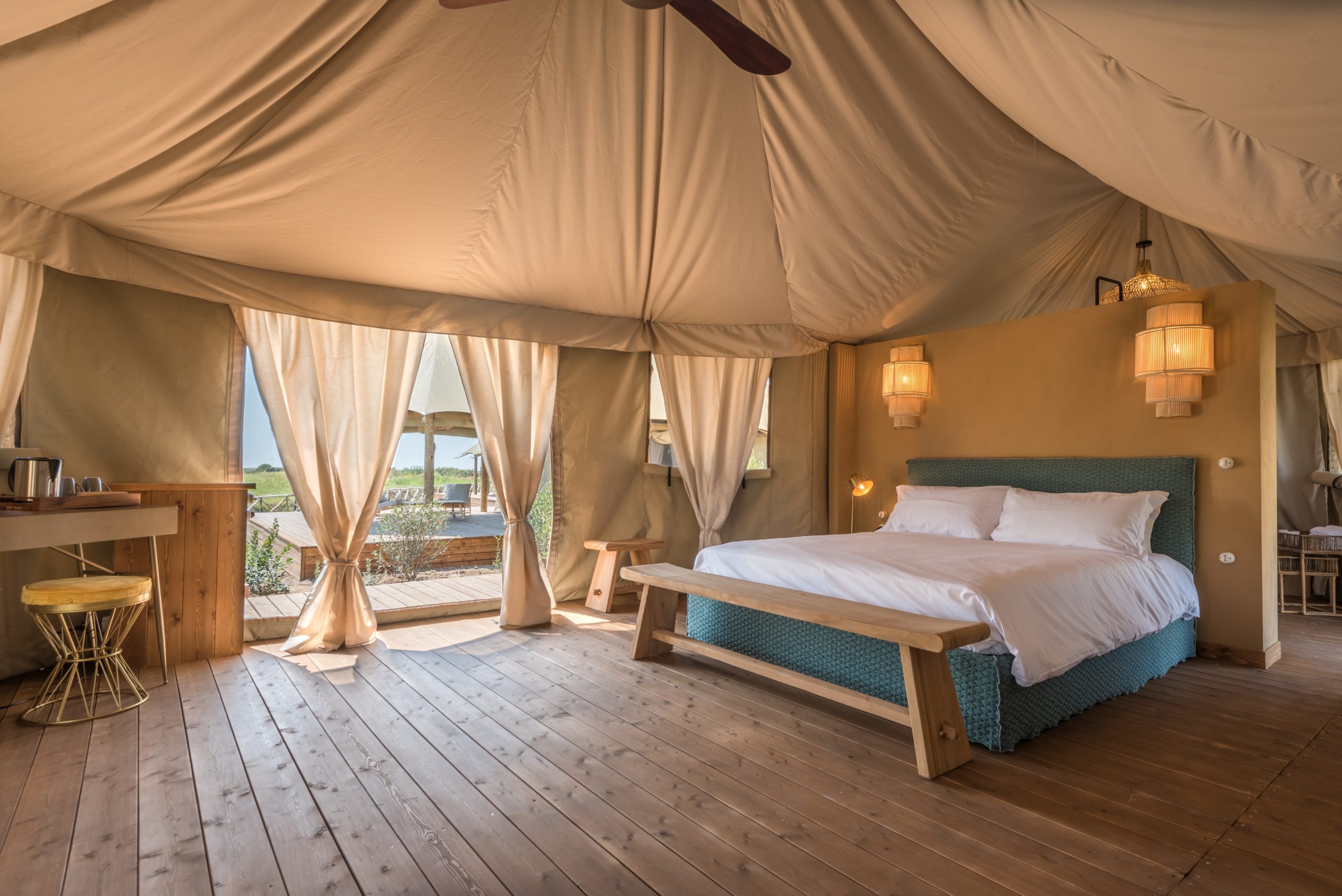 A tented bedroom with a wooden floor, white linen and warm woven lamps