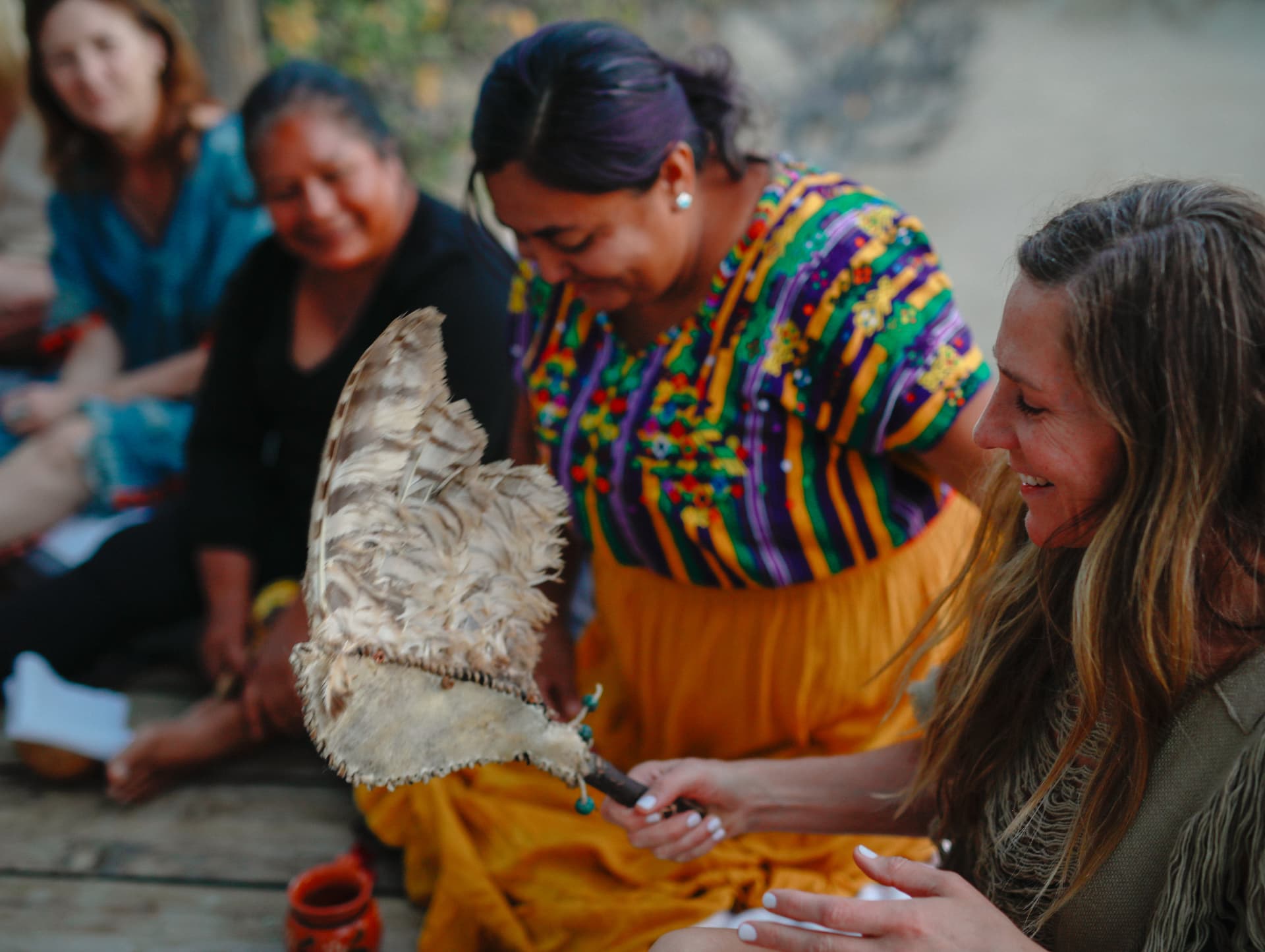 A woman smiling as she holds a feather fan beside women seated in circle