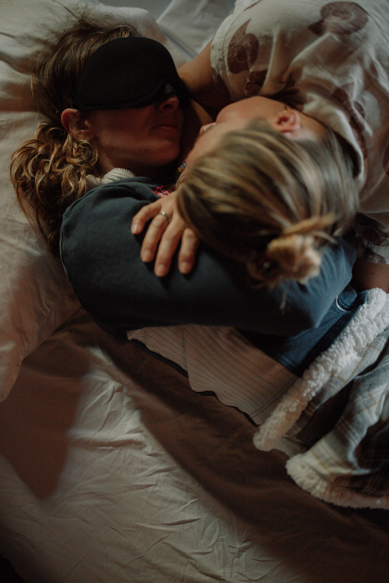 A woman wearing an eye mask sharing an embrace while resting beneath blankets