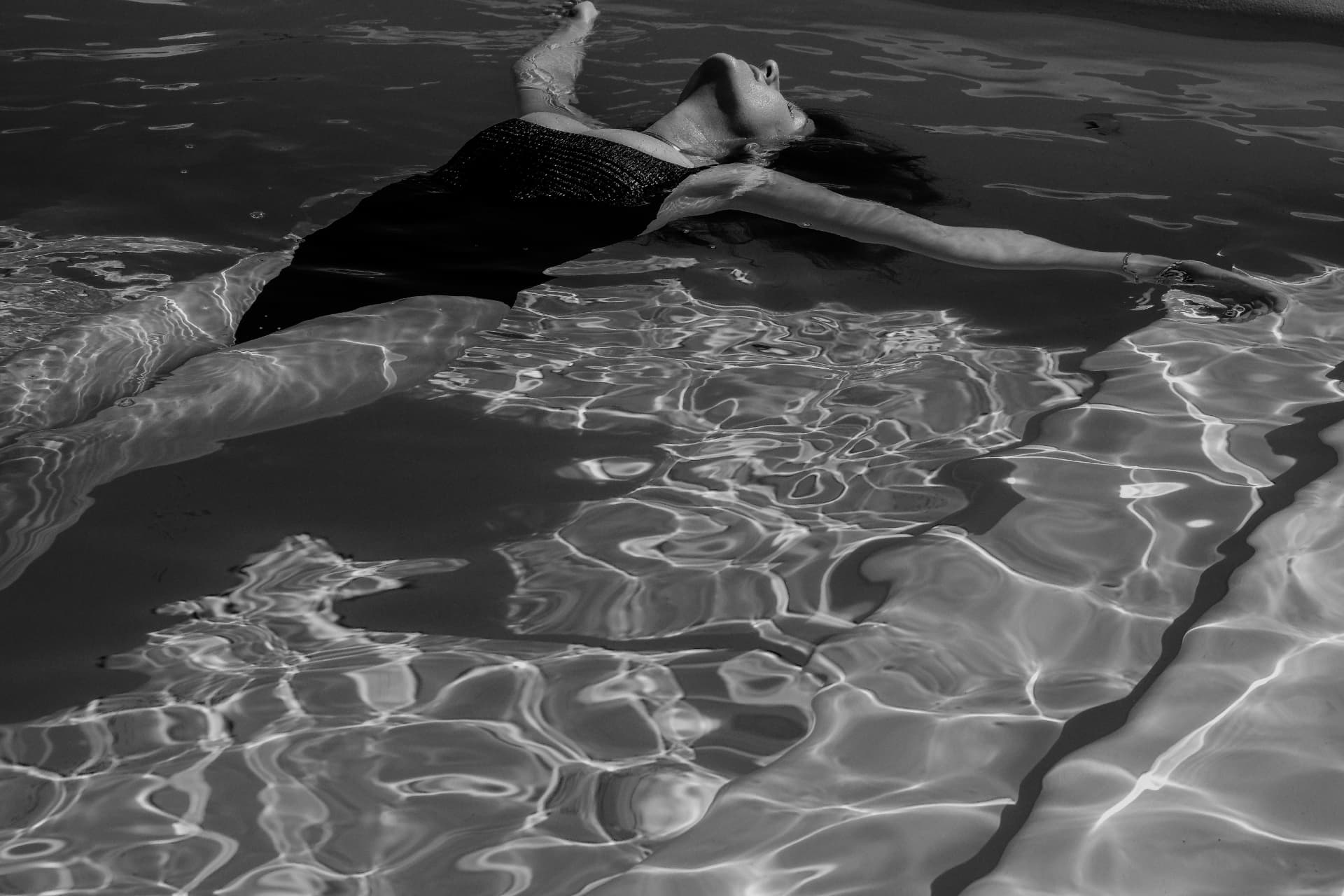 A woman floating in rippling pool water with her arms open, in black and white