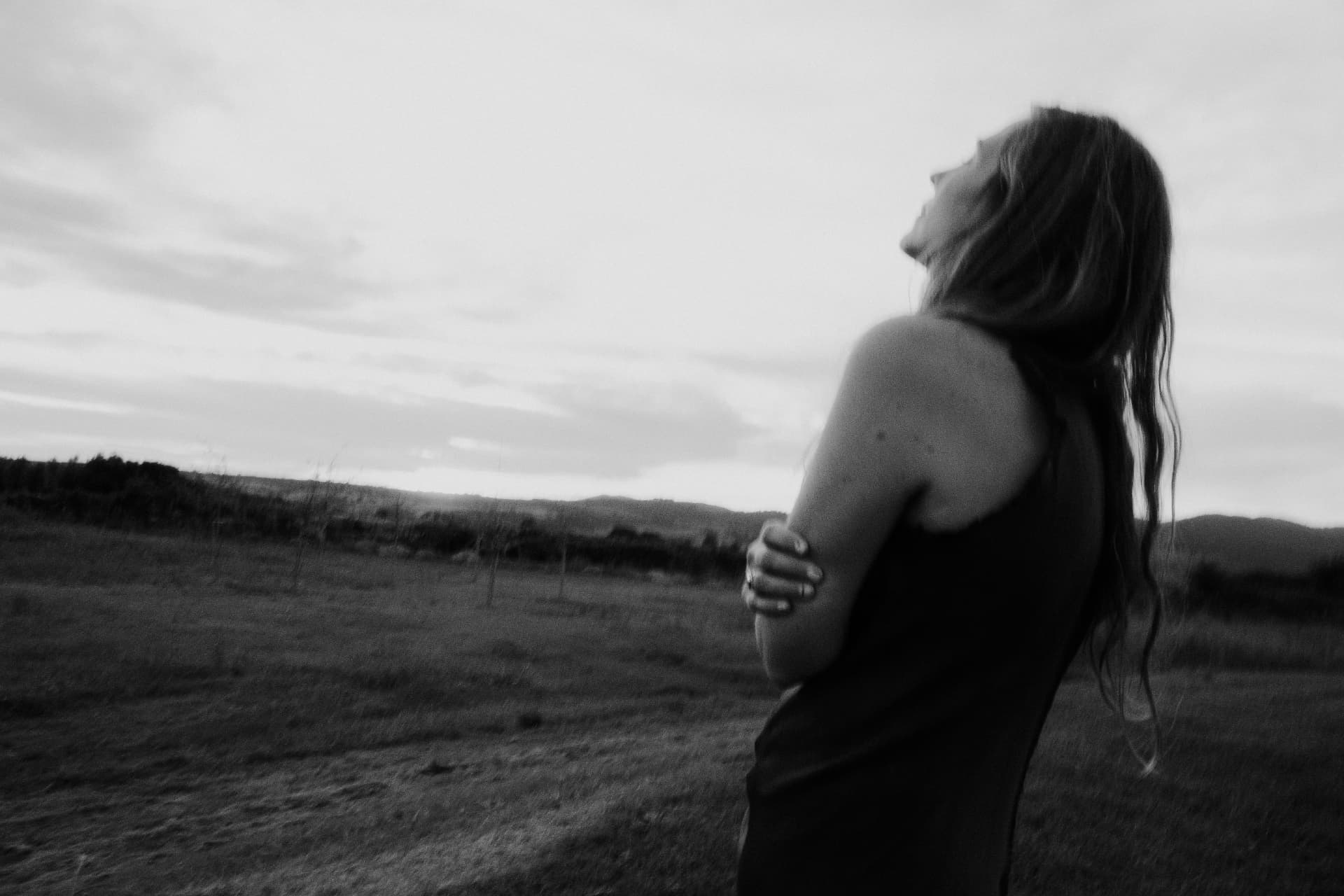 A woman holding herself and looking up at the sky over an open field, in black and white