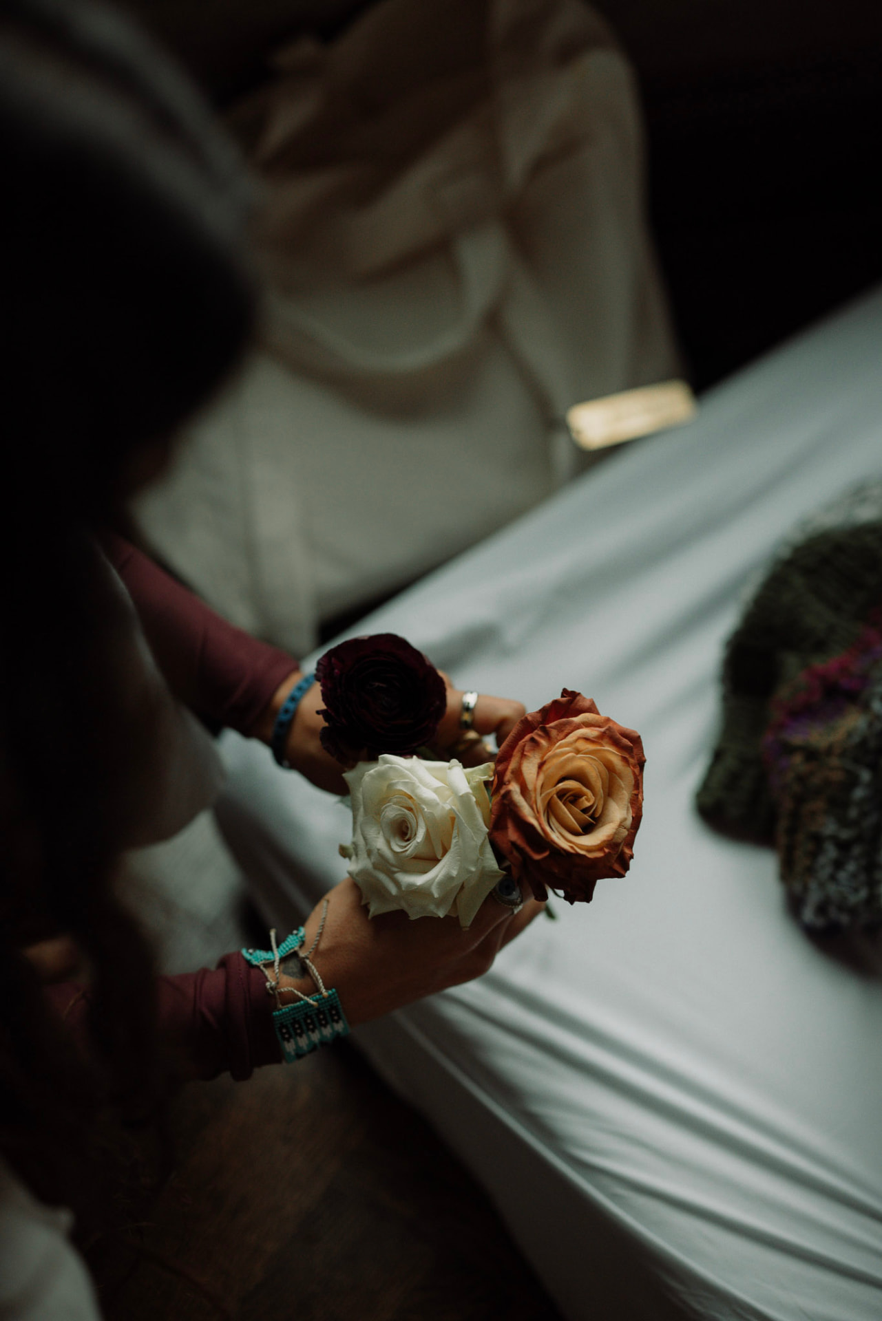 Hands holding a white rose, an apricot rose and a dark ranunculus above pale linen